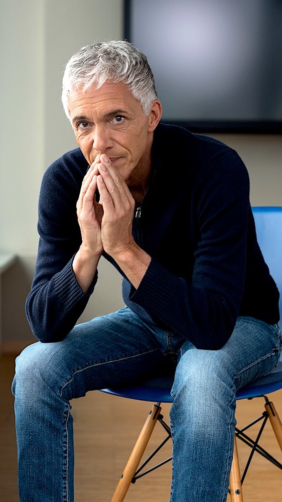 Michael C. Lauber auf eine blauen Stuhl in einem Konferenzzimmer. Trägt Casual Jeans und Pullover.
